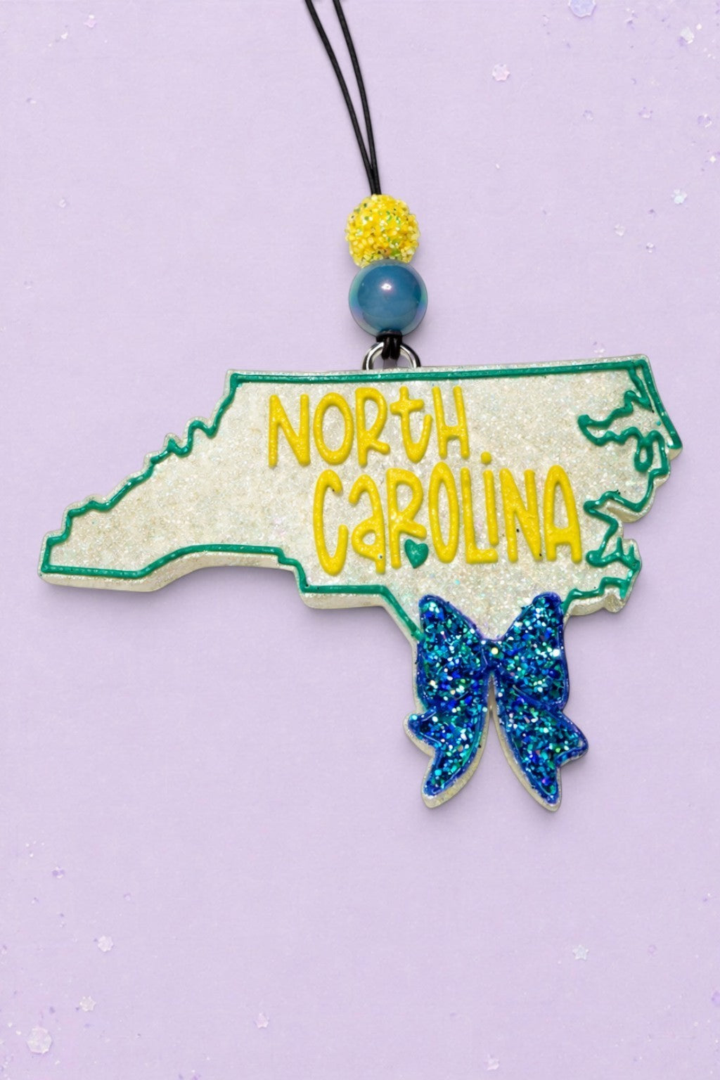 BeFreshie North Carolina state shaped car freshie with glittery white finish, yellow lettering, and a blue glitter bow.