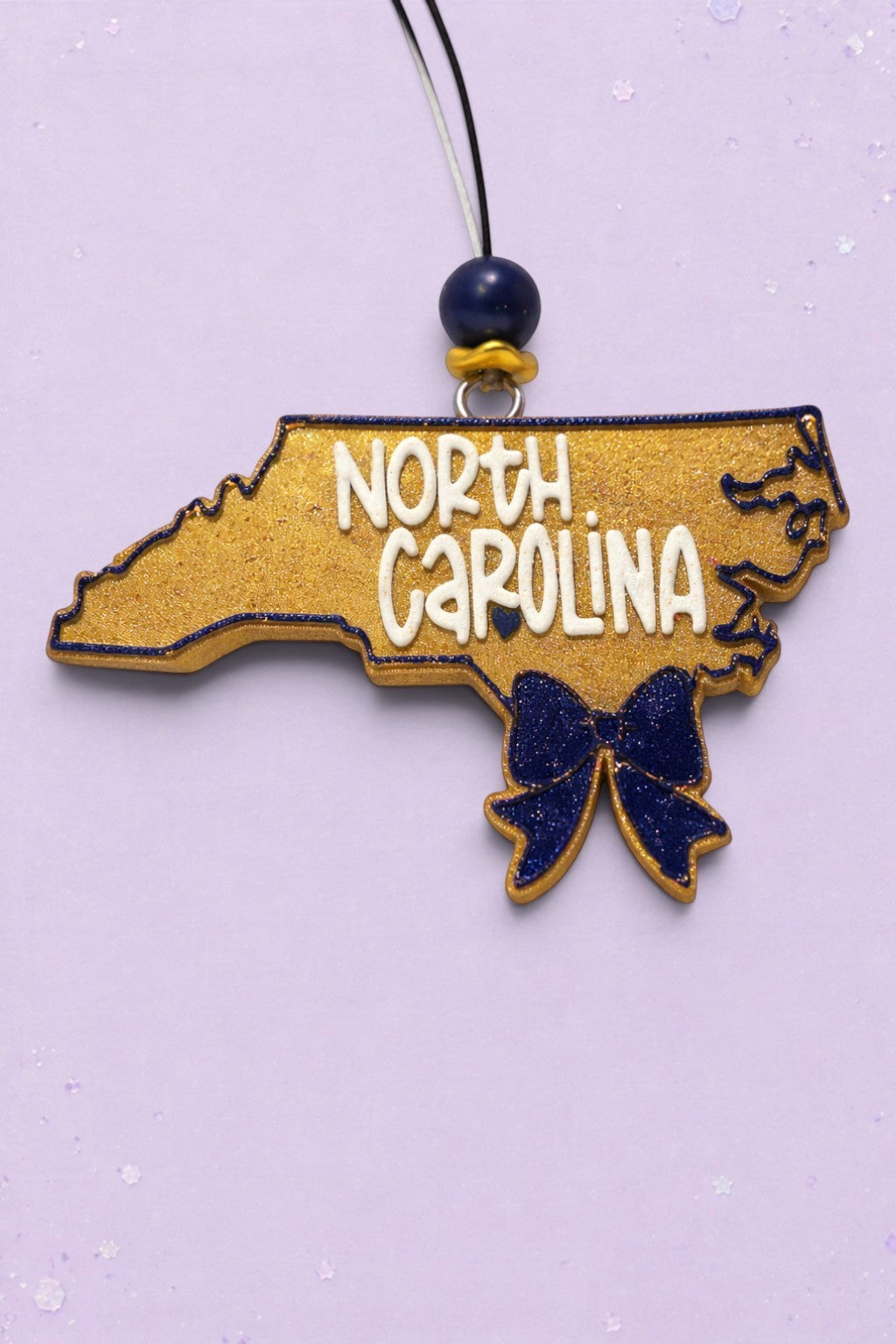 BeFreshie North Carolina state shaped car freshie in gold glitter with a navy blue bow and white lettering.